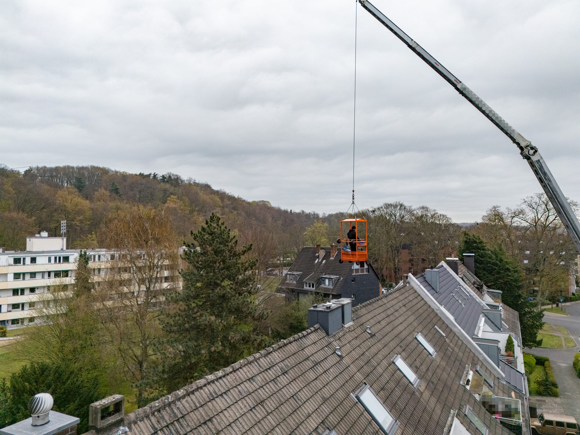 Drohnenaufnahme: Orangefarbener Arbeitskorb mit Person am Kranhaken über einem Ziegeldach
